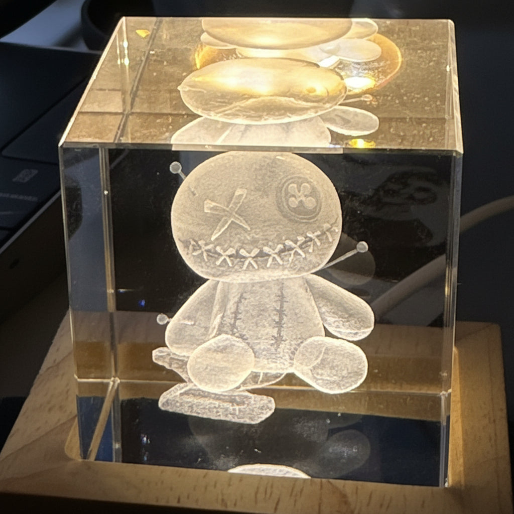 Clear crystal cube with an internally engraved design of a voodoo doll an anvil and a hammer.