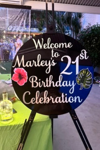 Round sign with 'Welcome to Marley's 21st Birthday Celebration' text on a stand, with a table and outdoor setting in the background.
