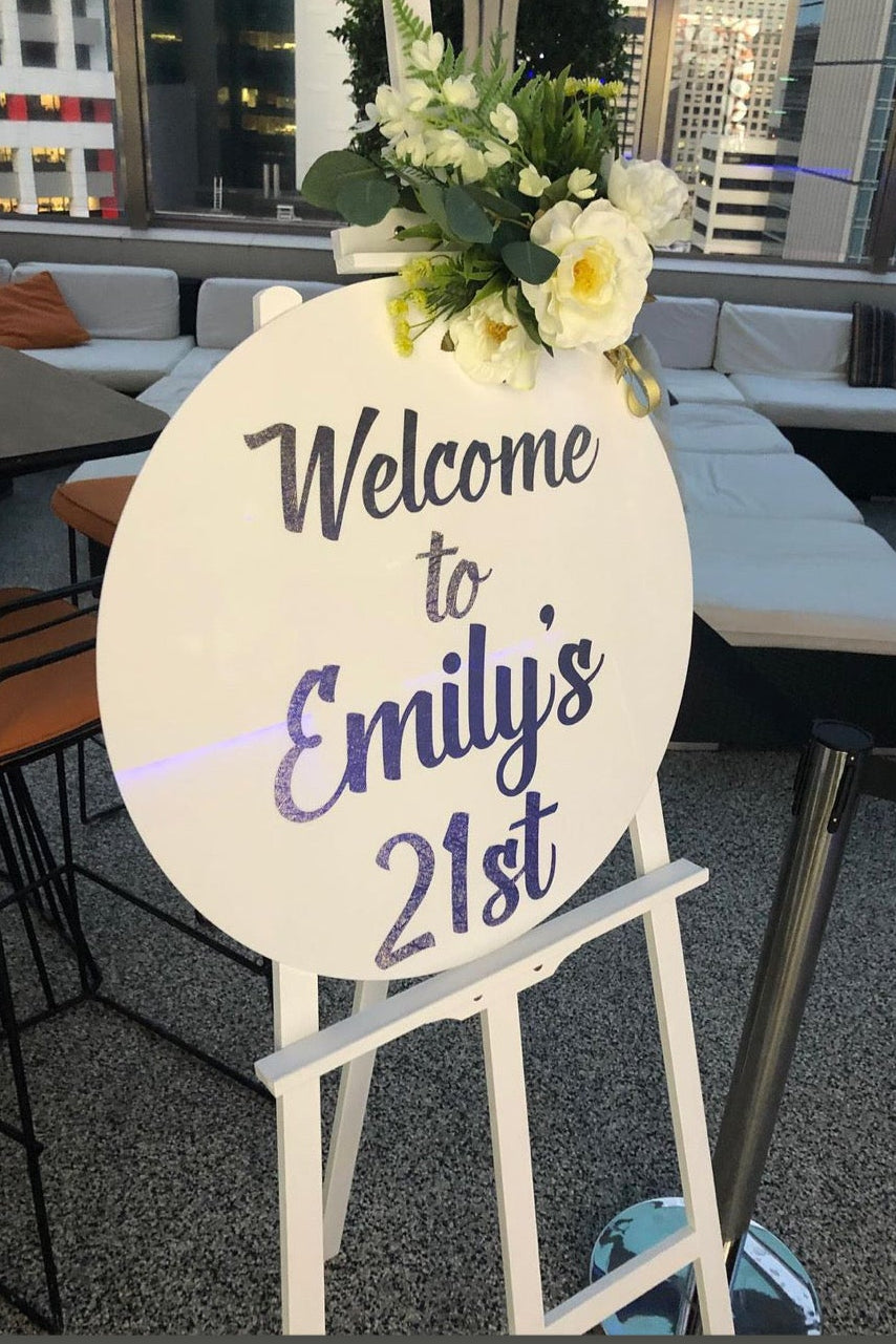 Decorative sign for Emily's 21st birthday with flowers on a stand in an indoor setting.